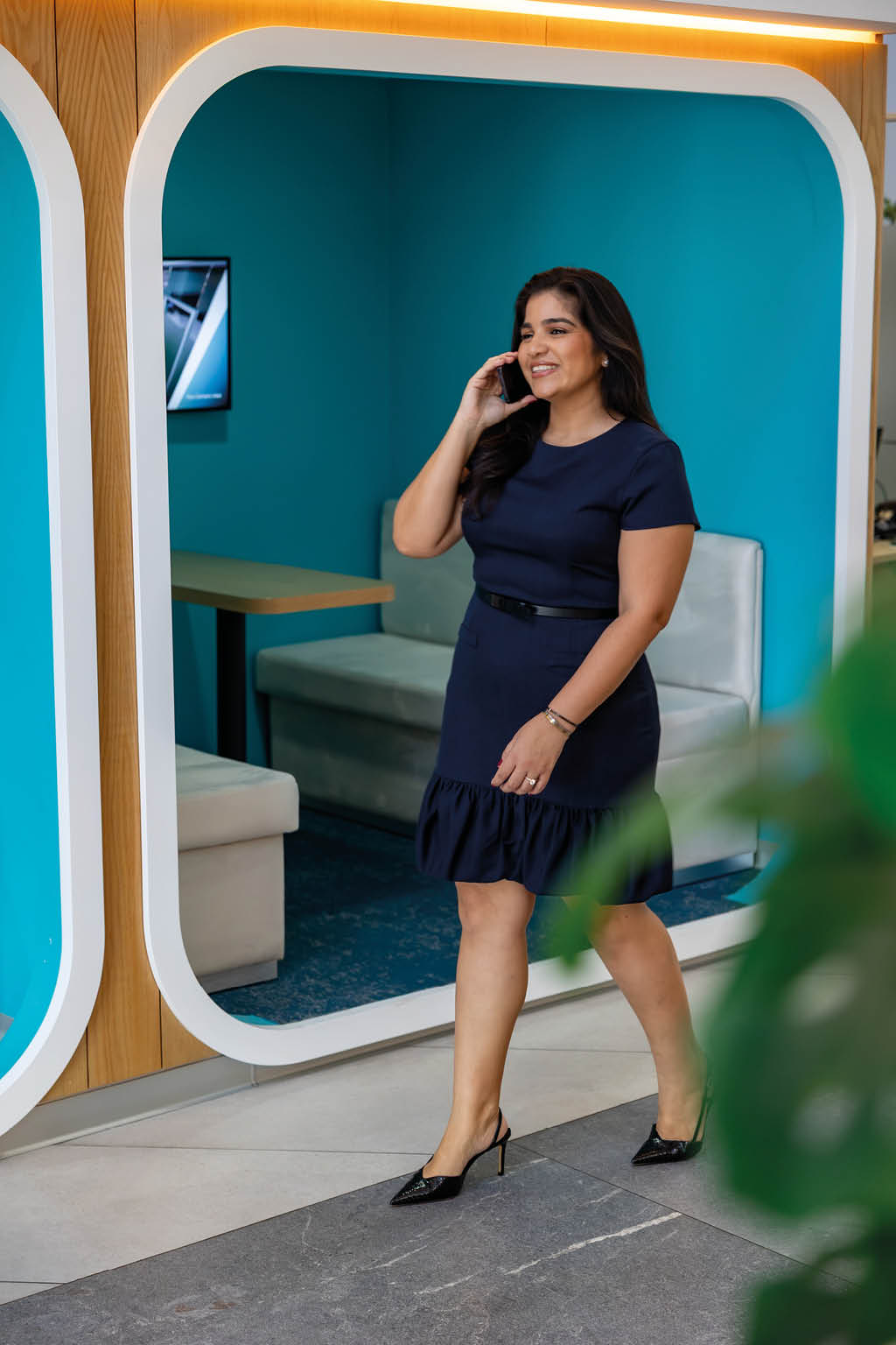 Persona con vestido azul oscuro y zapatos de tac n alto caminando y hablando por tel fono frente a una pared azul con un marco blanco y muebles tapizados. Descripci n generada con IA