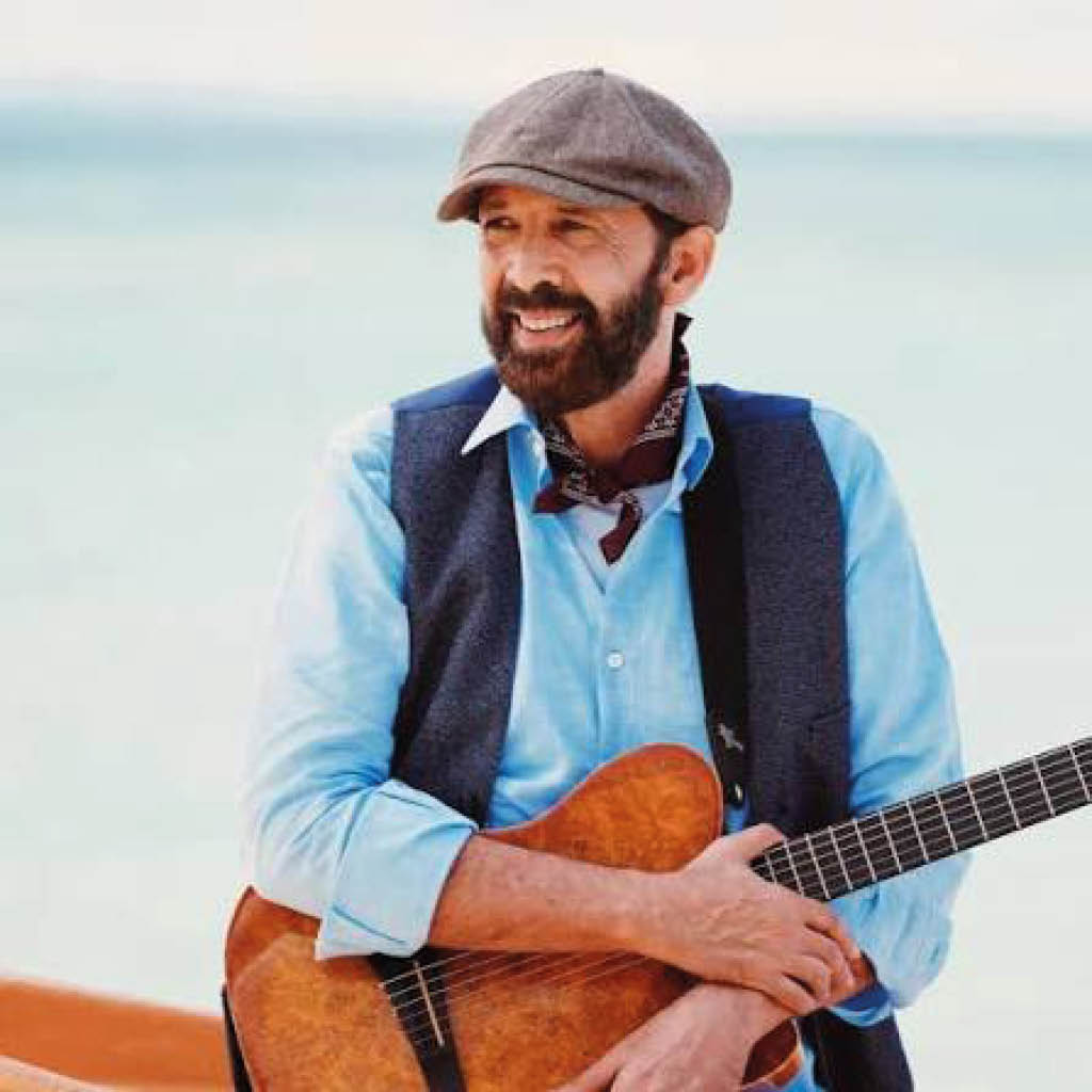 Una persona sonriente con barba y gorra est sentada junto al mar sosteniendo una guitarra ac stica mientras viste una camisa azul clara, chaleco oscuro y pa uelo al cuello.  Descripci n generada con IA