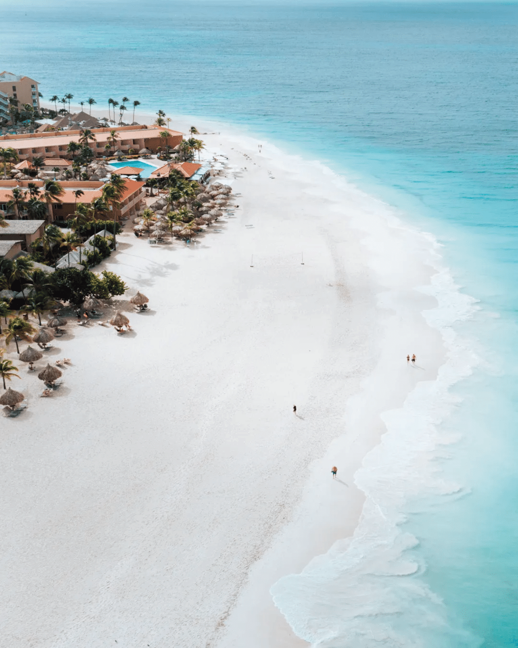 Playa amplia con arena blanca y agua turquesa junto a un complejo hotelero con palmeras y personas disfrutando del sol y el mar. Descripci n generada con IA