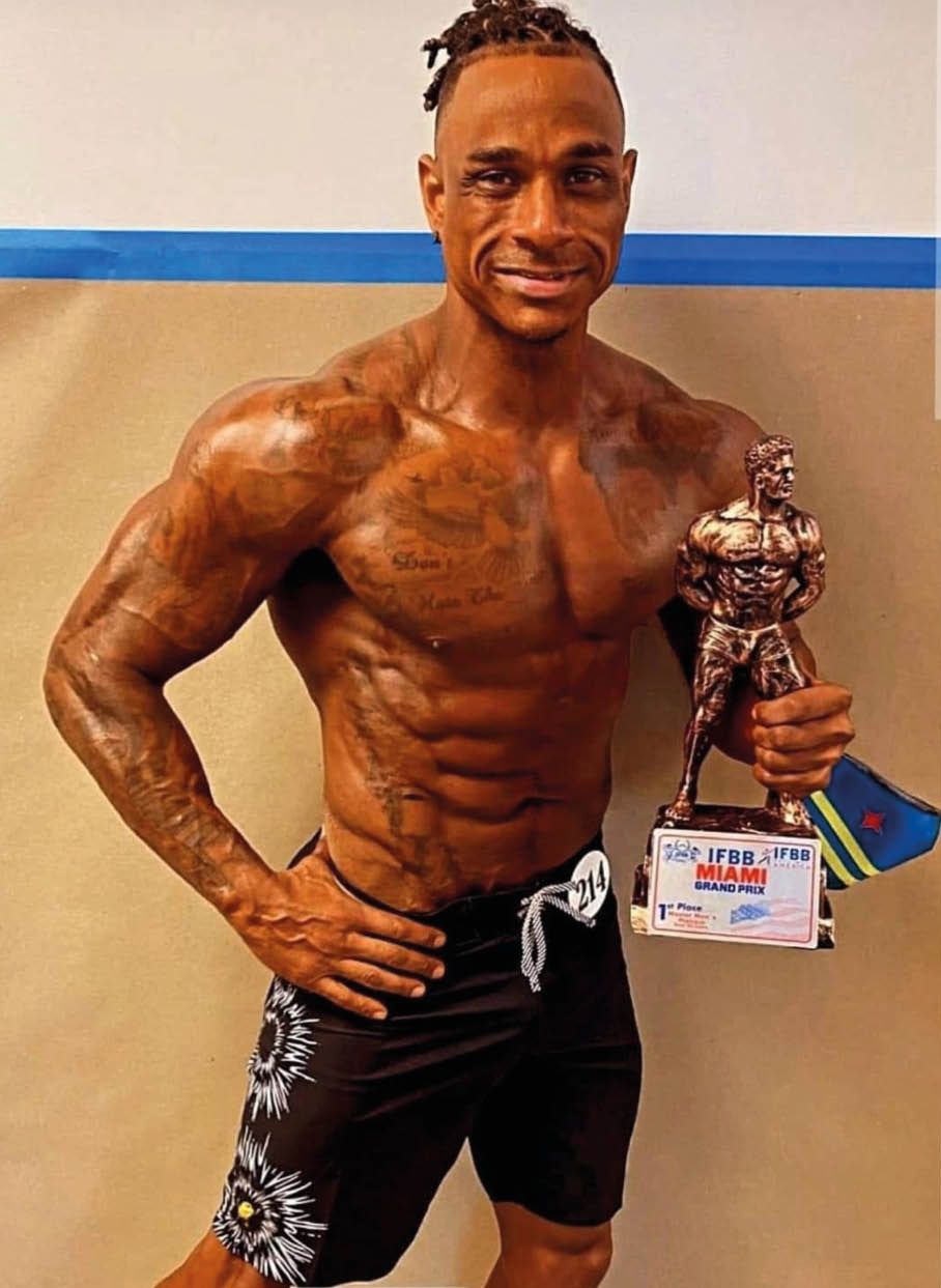 Persona con peinado de trenzas cortas y pantalones cortos negros con dise o floral sostiene un trofeo de culturismo y una placa de premio en un entorno interior. Descripci n generada con IA