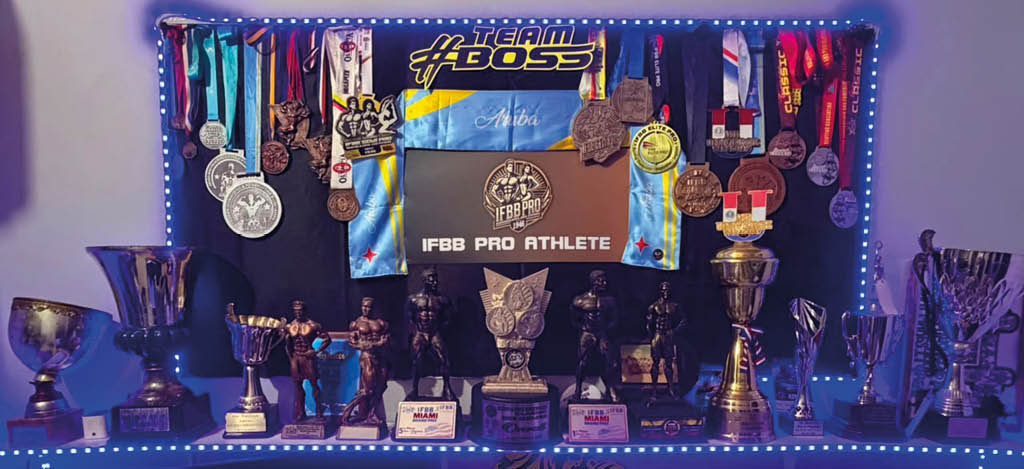 Una exhibici n iluminada con luces azules muestra m ltiples trofeos, medallas colgadas y figuras de personas en poses de culturismo con un cartel central que dice “IFBB Pro Athlete". Descripci n generada con IA