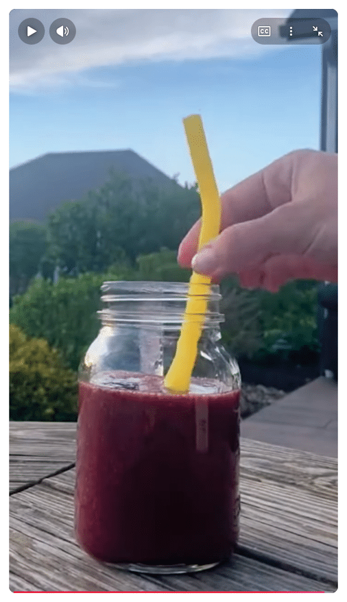 Un vaso de vidrio con un batido rojo oscuro sobre una mesa de madera al aire libre y una mano sosteniendo una pajilla amarilla flexible dentro del vaso. Descripci n generada con IA
