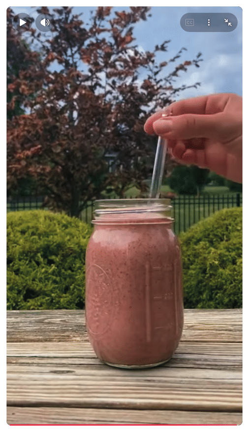 Un adulto sostiene un vaso de vidrio con un batido rosa y una pajilla mientras est al aire libre frente a arbustos y un  rbol.  Descripci n generada con IA