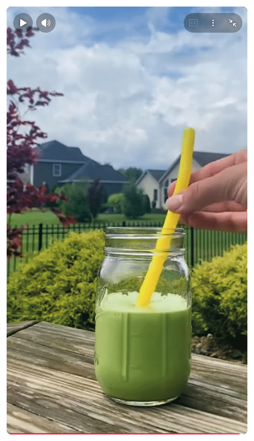 Un vaso de vidrio con bebida verde y una mano sosteniendo una pajilla amarilla al aire libre sobre una mesa de madera con un jard n y casas al fondo. Descripci n generada con IA