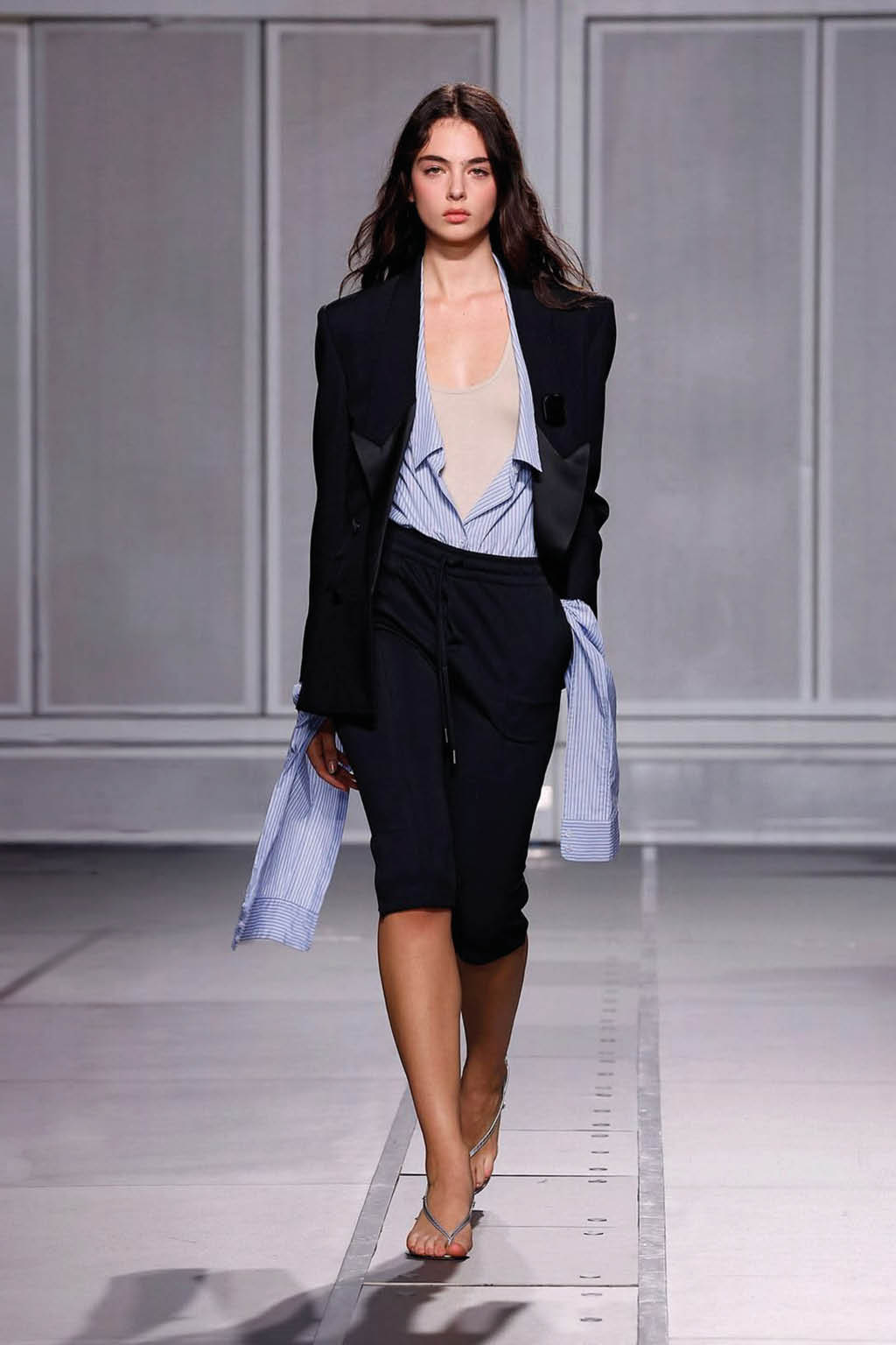 Una persona con cabello largo y suelto camina por una pasarela vistiendo una chaqueta negra, una camisa azul de rayas parcialmente desabotonada y pantalones cortos negros, con sandalias. Descripci n generada con IA
