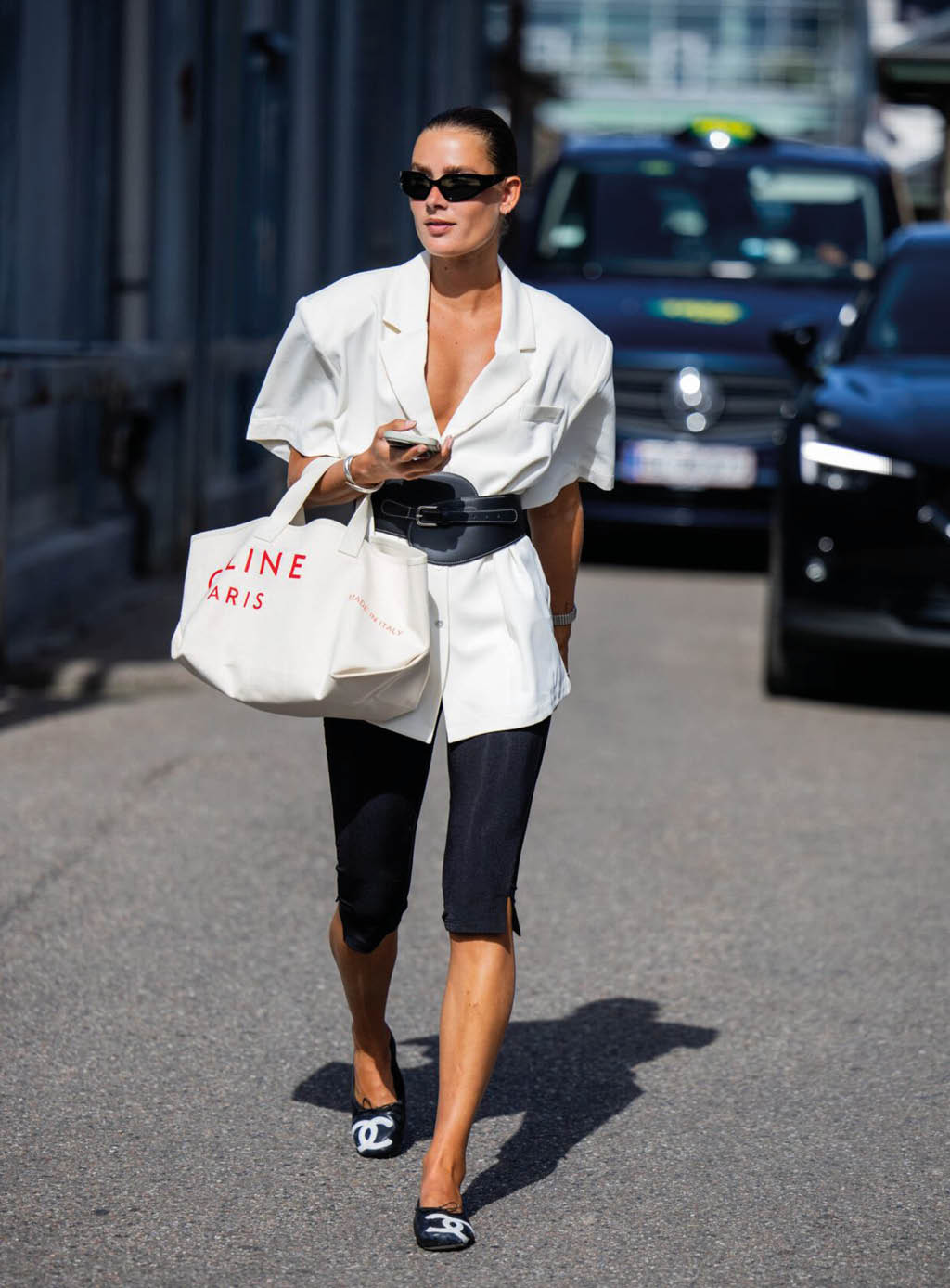 Una persona con gafas de sol lleva una chaqueta blanca con cintur n negro, pantalones cortos negros y zapatos con el logo de Chanel mientras camina por una calle urbana con un bolso blanco grande. Descripci n generada con IA