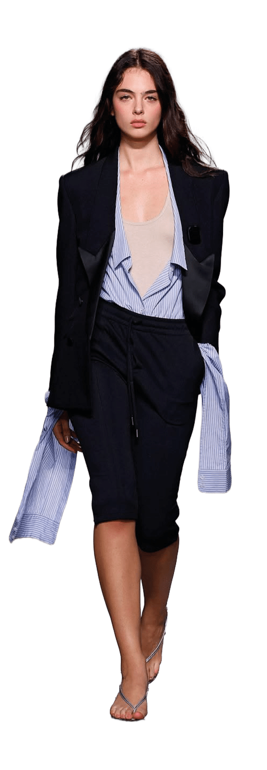 Una persona con cabello largo y suelto camina con confianza vistiendo un conjunto elegante que combina una chaqueta negra, una blusa de rayas azul y blanca y pantalones cortos negros, complementado con sandalias. Descripci n generada con IA