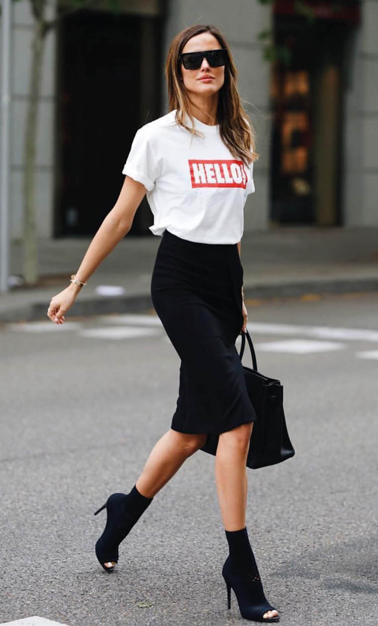 Una persona con gafas de sol, camiseta blanca con la palabra “HELLO" en rojo, falda negra ajustada y zapatos de tac n altos camina por una calle urbana mientras lleva un bolso negro. Descripci n generada con IA