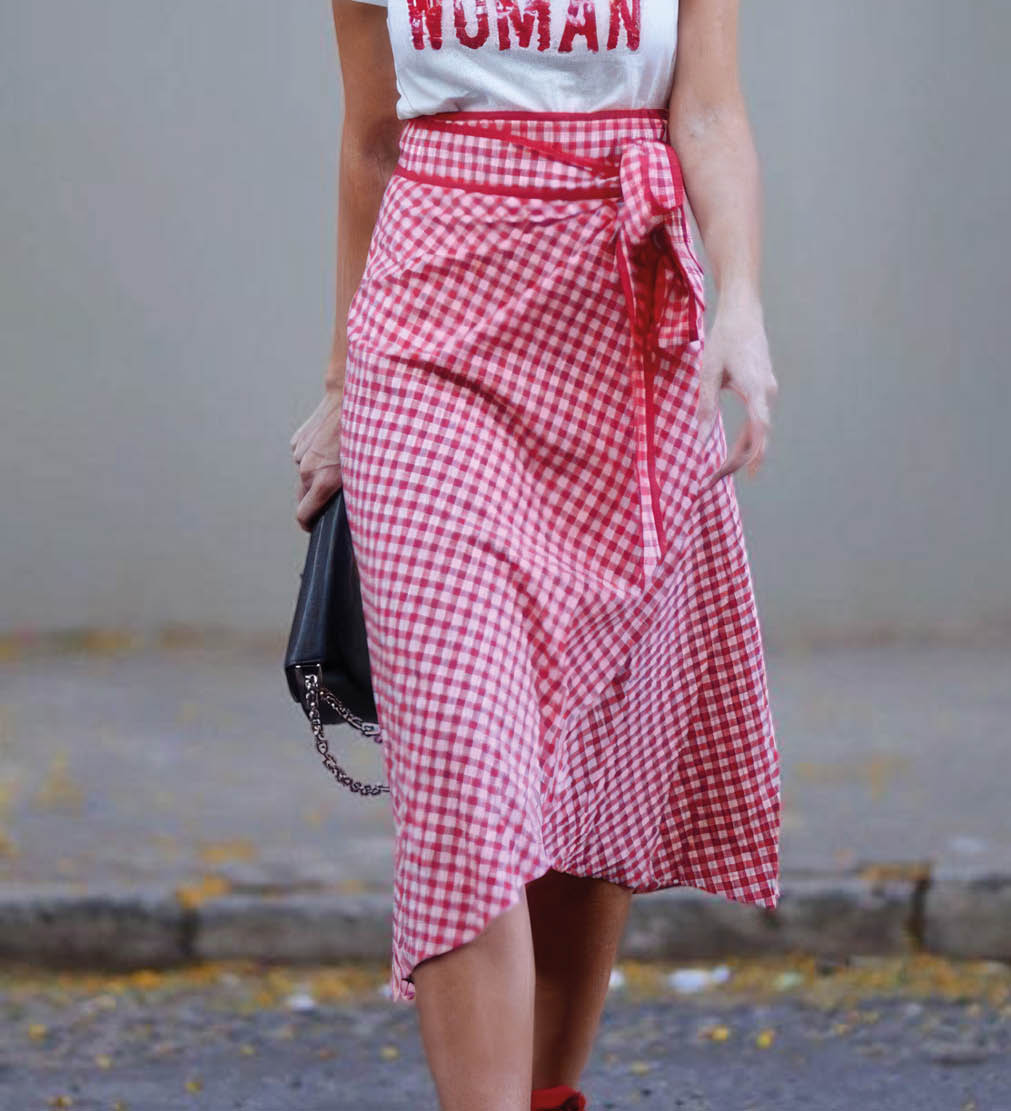 Persona con cabello largo y ondulado que lleva gafas de sol, camiseta blanca con texto rojo, falda larga de cuadros rojos y sandalias rojas caminando por una calle. Descripci n generada con IA