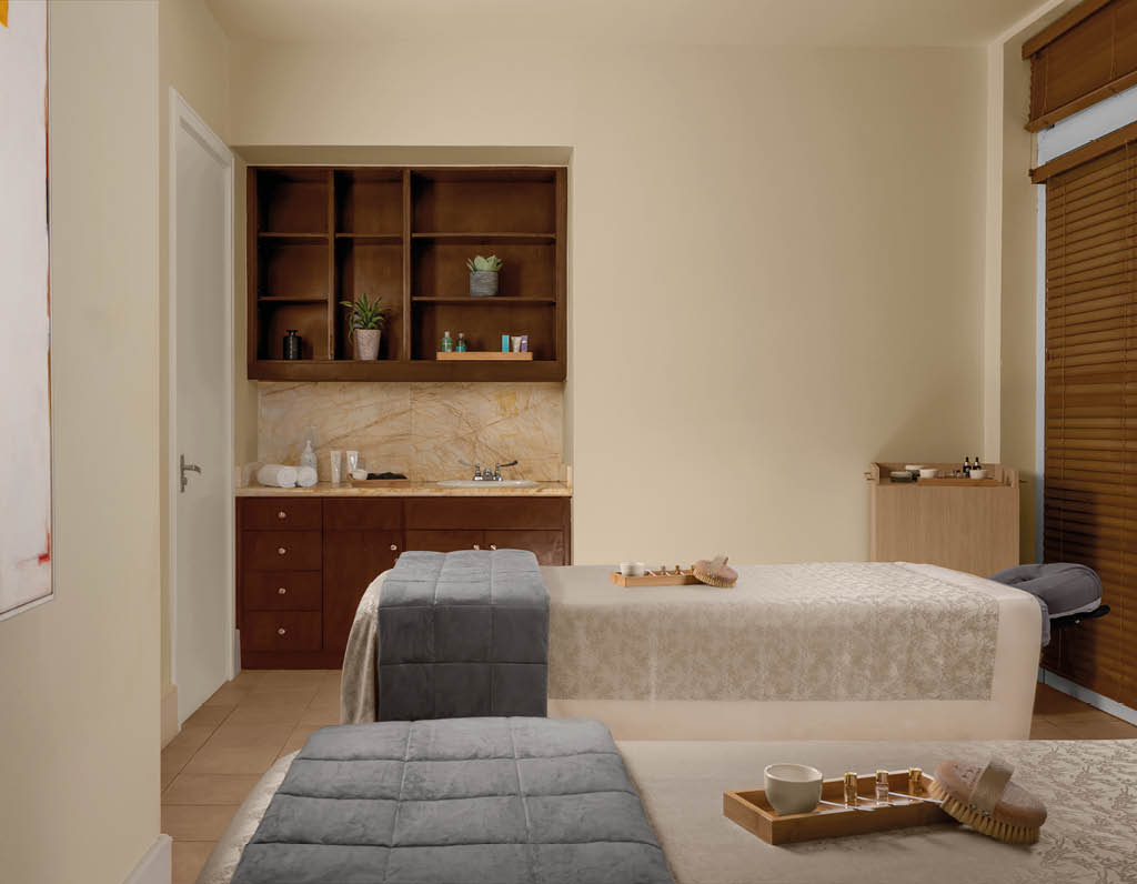 Habitaci n con dos camillas cubiertas con mantas beige y grises, una pared con estanter as de madera y una ventana con persianas de madera. Descripci n generada con IA