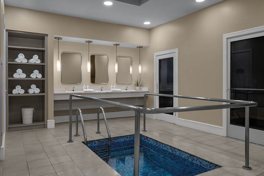 Sala de hidroterapia con piscina peque a, toallas blancas dobladas en estantes, espejos y lavabos con dispensadores, y paredes beige con puertas de vidrio. Descripci n generada con IA