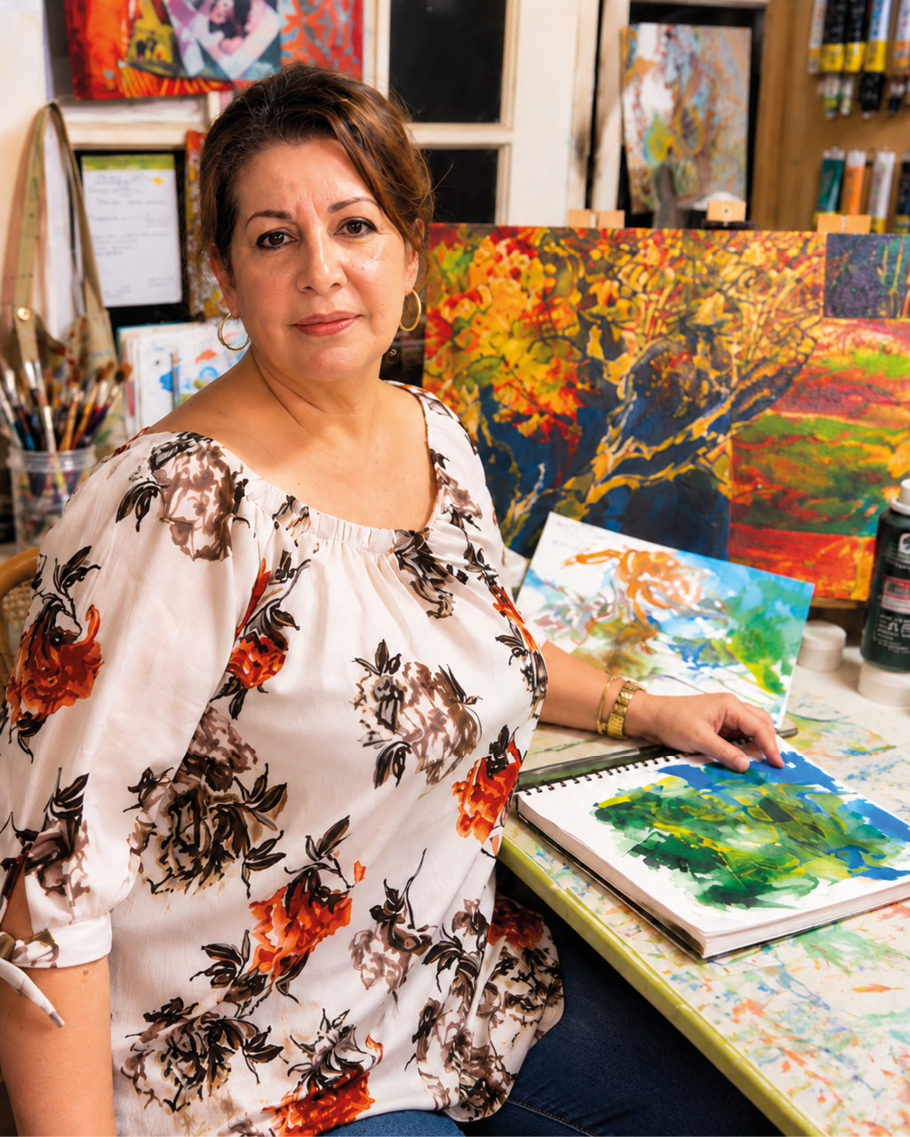Persona con cabello recogido y blusa blanca con estampado floral naranja y marr n sentada en un estudio de arte mientras sostiene un pincel y trabaja en una pintura colorida sobre una mesa llena de materiales art sticos. Descripci n generada con IA