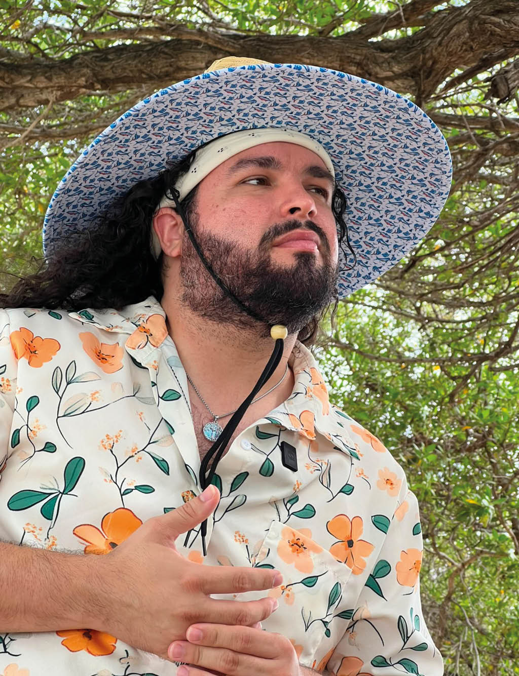 Persona con cabello largo y barba que lleva un sombrero amplio y una camisa blanca con estampado floral naranja y verde, posando al aire libre bajo un rbol.  Descripci n generada con IA