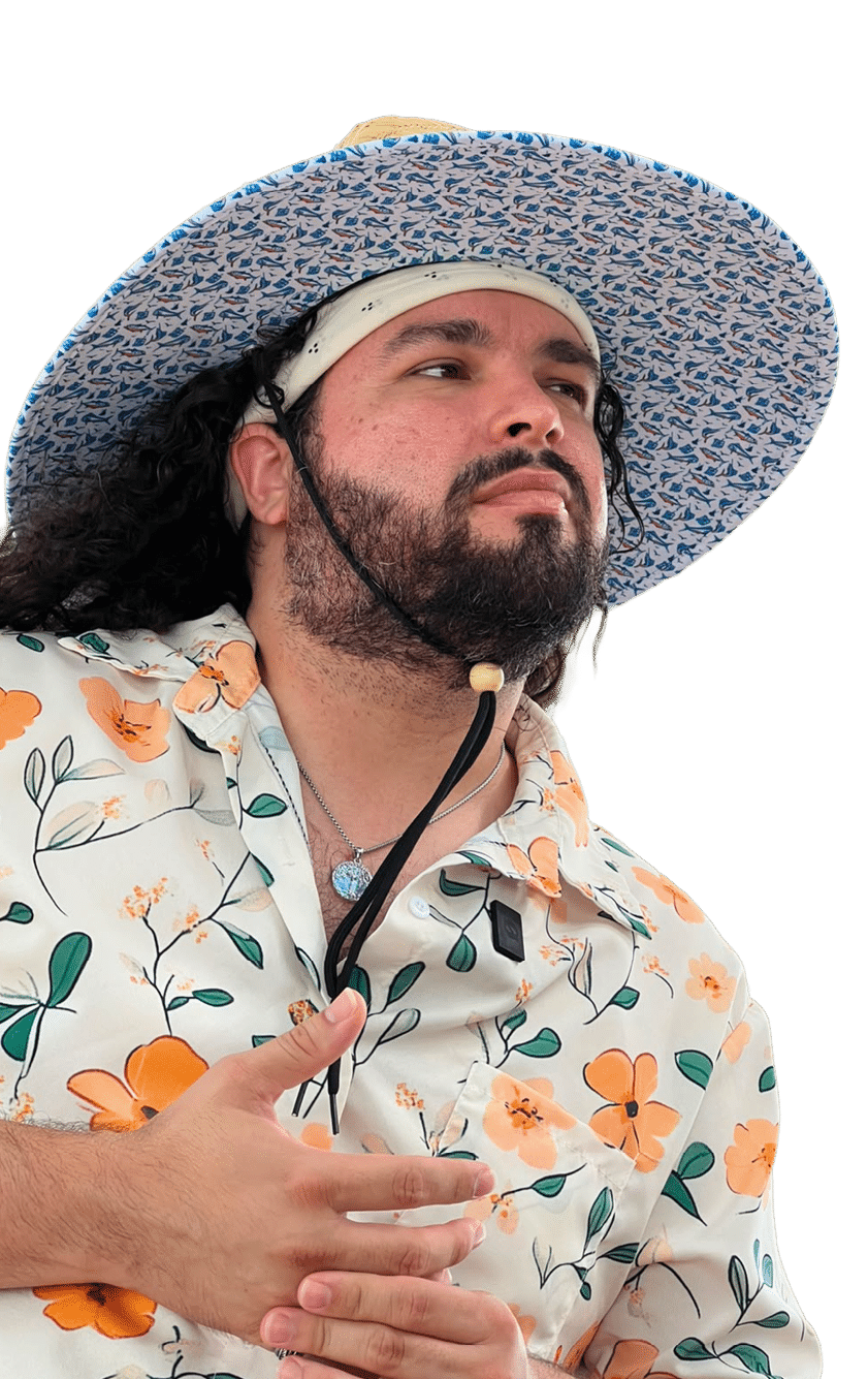 Persona con barba y cabello largo que lleva un sombrero de ala ancha con estampado azul y una camisa blanca con flores naranjas y verdes mientras mira hacia un lado. Descripci n generada con IA