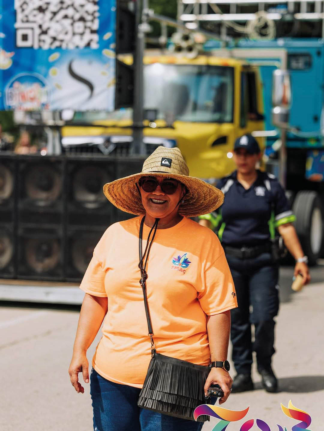 Una persona con sombrero de paja, gafas de sol y camiseta naranja sonr e mientras sostiene un bolso negro y camina en un evento al aire libre con otra persona uniformada al fondo. Descripci n generada con IA