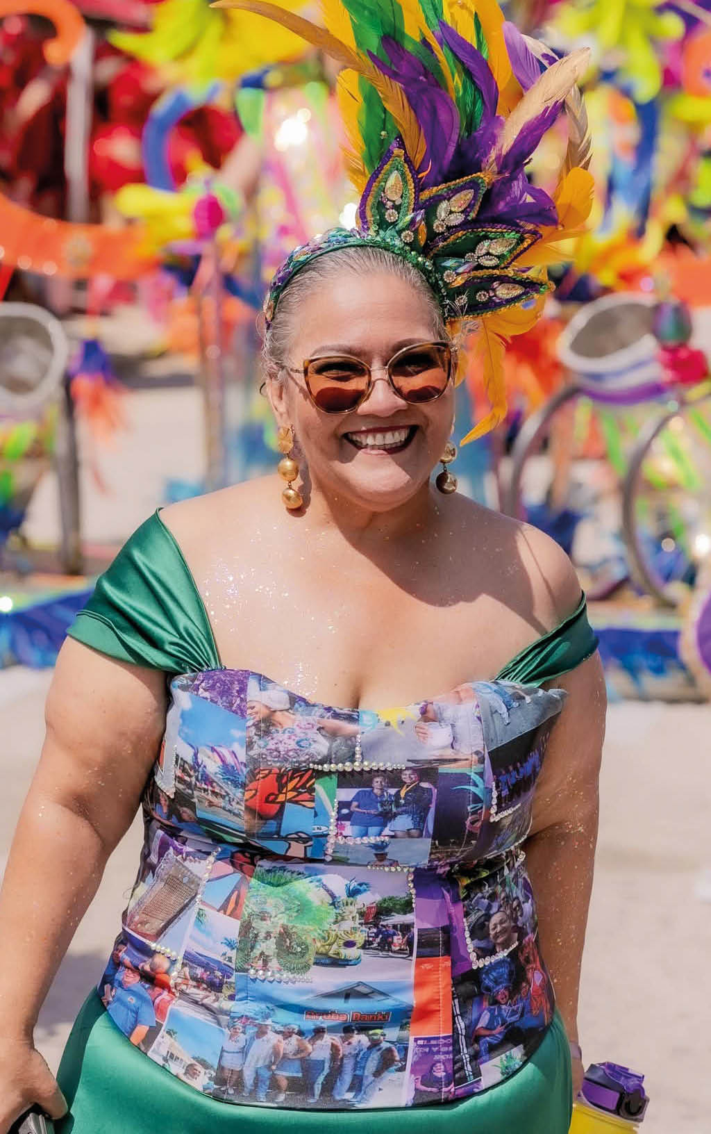 Persona sonriente con vestido verde y cors colorido con estampados, usando gafas de sol y un tocado con plumas verdes, moradas y amarillas en un ambiente festivo y colorido.  Descripci n generada con IA