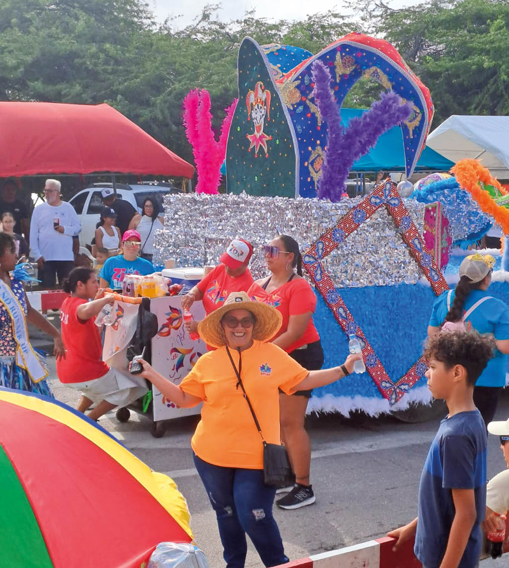 Una persona con sombrero de paja y camiseta naranja sonr e mientras sostiene un tel fono frente a un grupo de personas alrededor de una carroza decorada con colores brillantes y plumas en un evento al aire libre. Descripci n generada con IA
