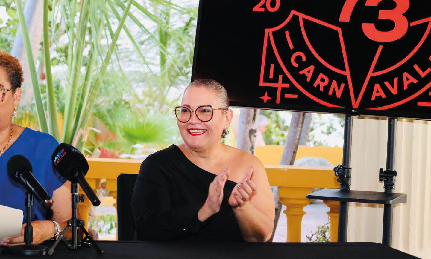 Persona con gafas y blusa negra de un solo hombro aplaudiendo mientras est sentada en una mesa con micr fonos frente a un fondo con plantas y un cartel negro con texto rojo relacionado con un carnaval.  Descripci n generada con IA