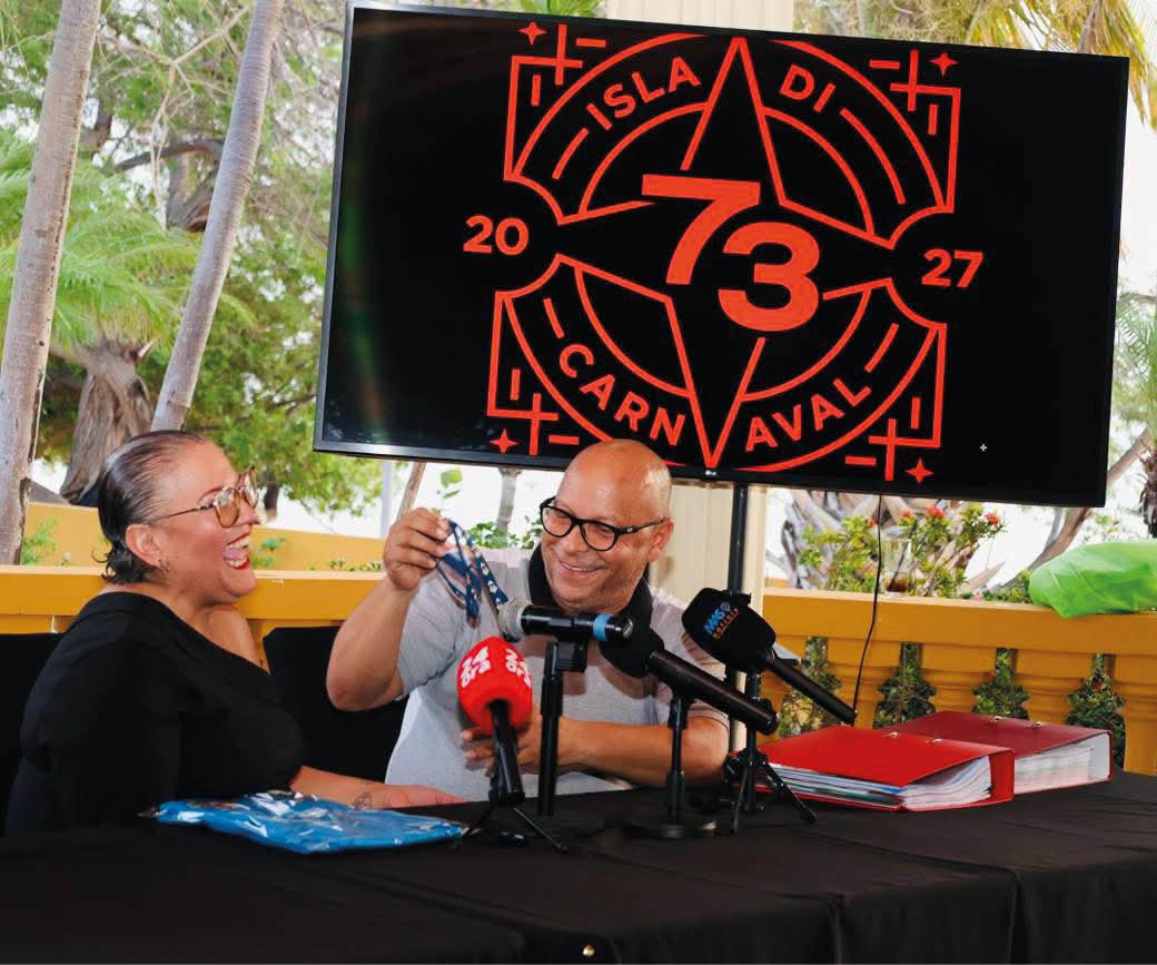 Dos personas sonrientes sentadas en una mesa con micr fonos frente a ellas mientras una pantalla muestra un logo de carnaval con texto y n meros. Descripci n generada con IA
