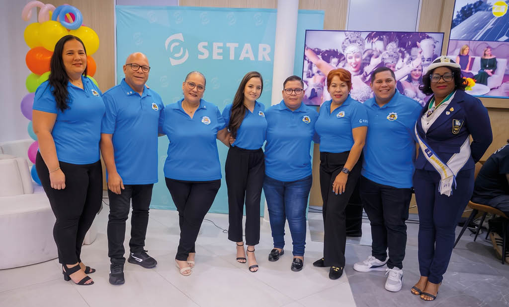 Ocho personas vestidas con camisetas azules posan sonrientes en un espacio interior decorado con globos y un cartel que dice SETAR. Descripci n generada con IA